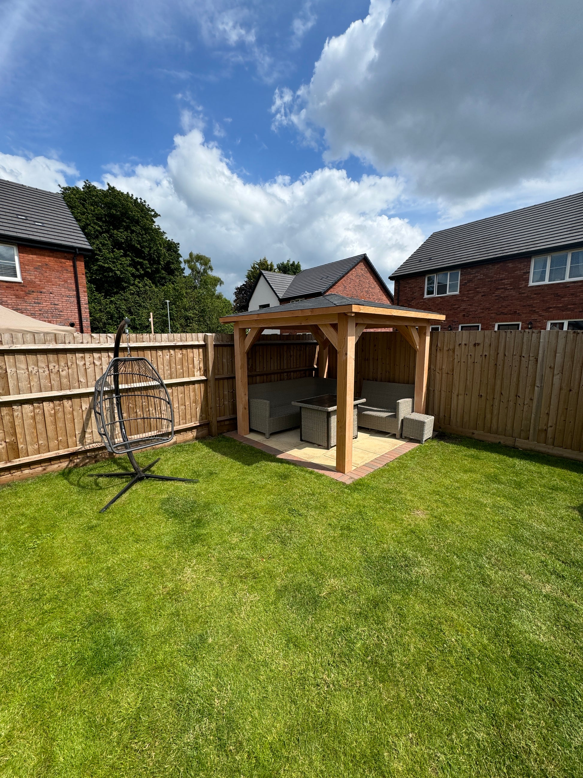 Patio area with a wooden gazebo with outdoor furniture on a grassy lawn under a blue sky.