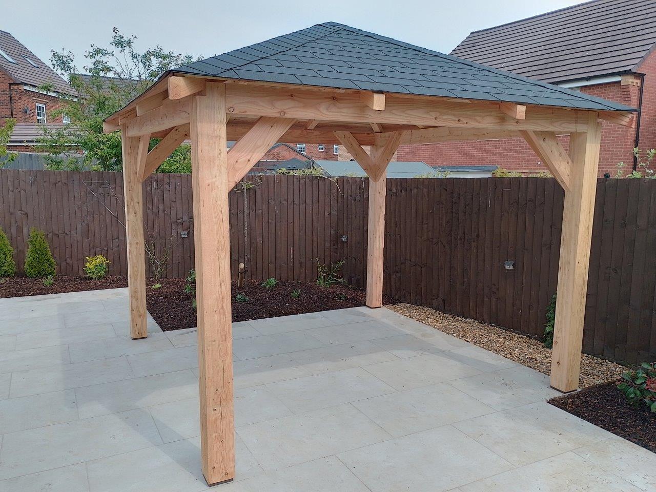Wooden gazebo with a black felt shingle roof on a patio area with a garden and houses in the background.
