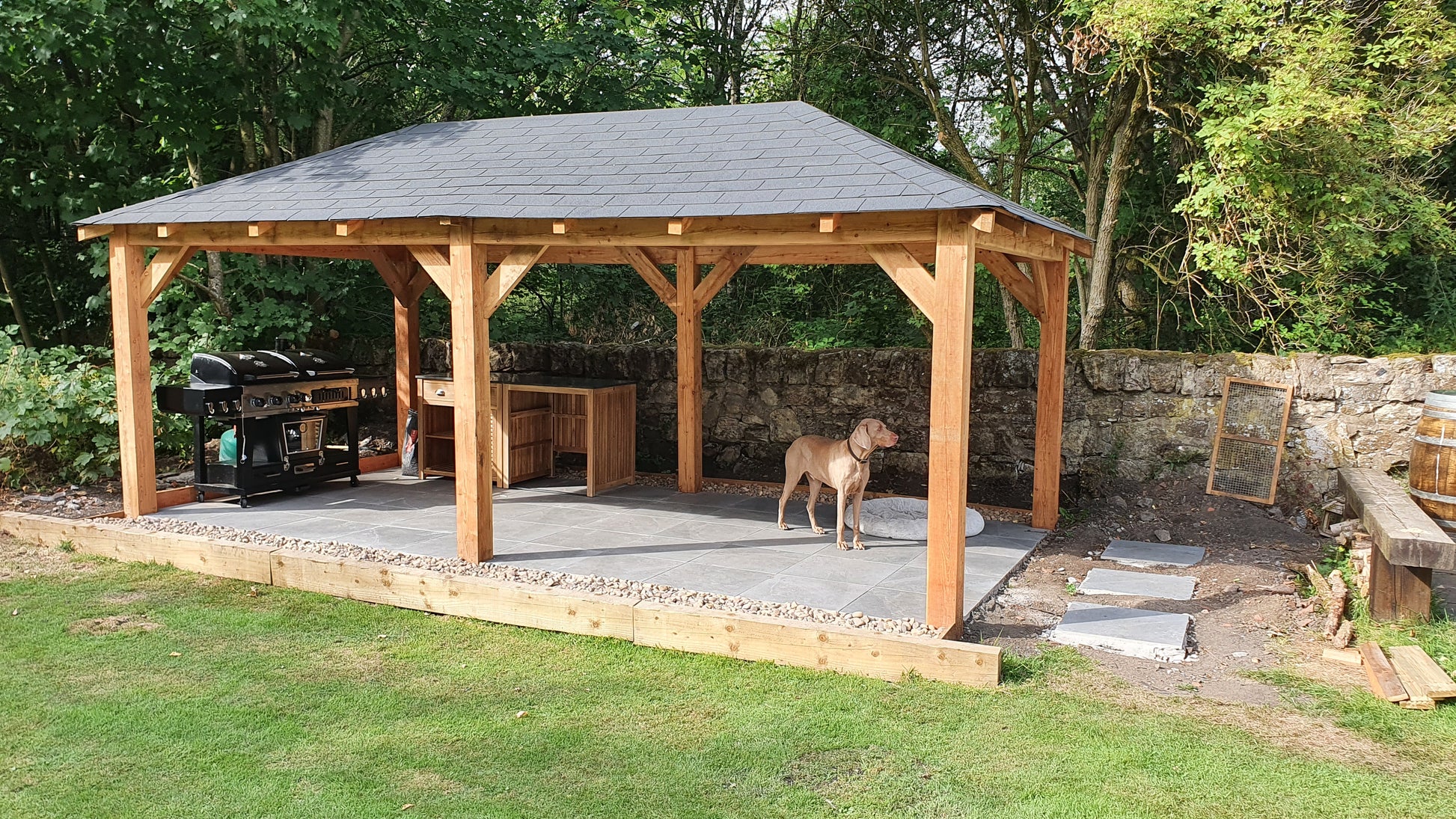 Wooden gazebo with a roof in a garden setting with a dog standing inside.